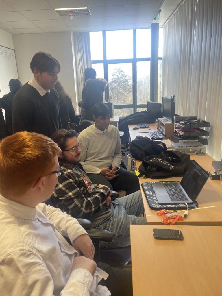 Three students sit around a laptop with our Head of Station Sound, Freddie Lee. The students are wearing white shirts and are sat in front of a large window in the Media Suite.